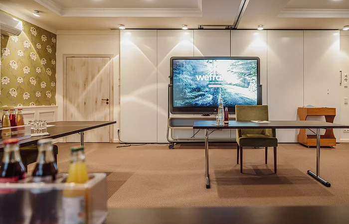 Konferenzraum mit einem großen Bildschirm, einem Tisch mit Getränken und Stühlen, florale Tapete an der Wand.