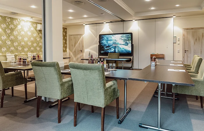 Konferenzraum mit grünen Stühlen, Tischen, Getränken und einem großen Bildschirm an der Wand.