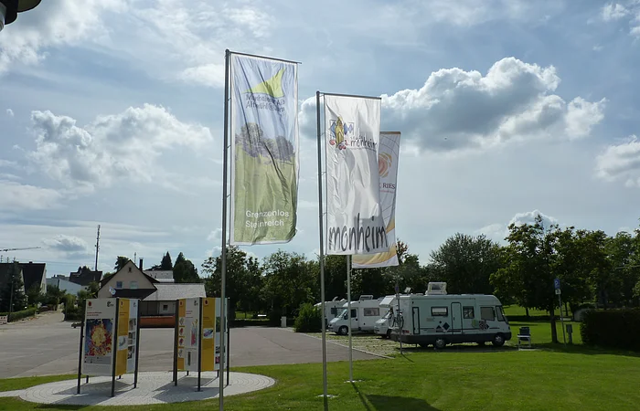 Drei Fahnen wehen im Wind, daneben stehen Informationstafeln auf einer Wiese. Im Hintergrund sind Wohnmobile und Bäume zu sehen.