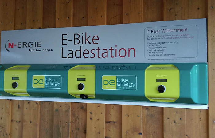 E-Bike-Ladestation mit drei Ladepunkten an einer Holzwand. Schild mit Logo und Informationen.