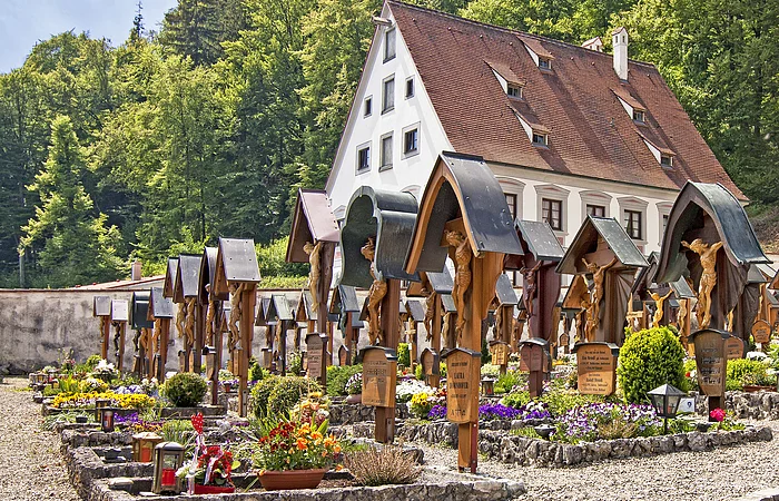 Ein Friedhof mit Holzkreuzen und Blumen, im Hintergrund ein Gebäude mit rotem Dach und Bäumen.