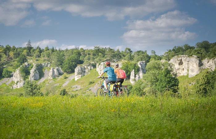 Zwei Personen fahren mit Fahrrädern auf einem grasbewachsenen Feld vor einer Felslandschaft.