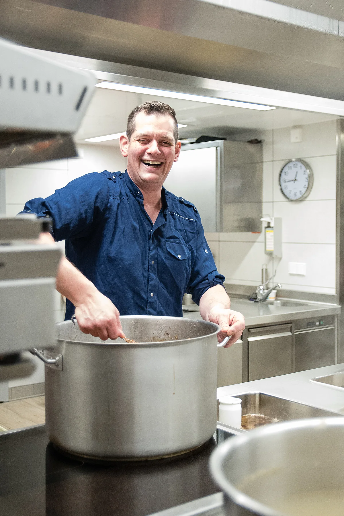 Andreas Blob lacht freudestrahlend und rührt in einem riesigen silbernen Kochtopf. Die restliche Küche ist leer, mit weißen Fliesen und silbernen Arbeitsplatten ausgestattet.