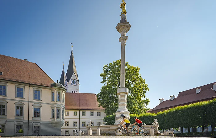 Zwei Radfahrer fahren an einer Mariensäule auf einem Platz mit historischen Gebäuden vorbei.