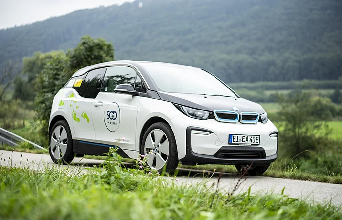 Ein weißes Elektroauto mit SGD Pharma-Logo fährt auf einer ländlichen Straße, im Hintergrund sind Bäume und Hügel.