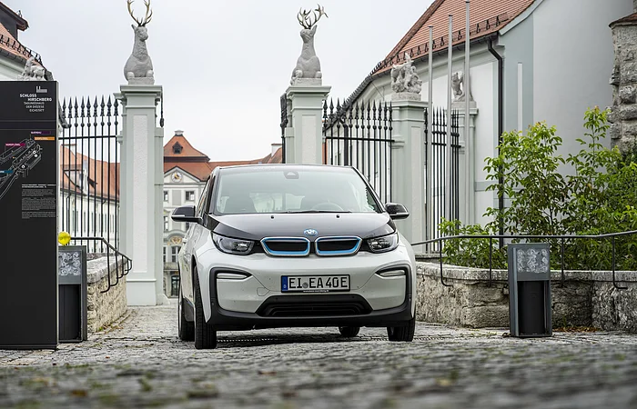 Ein Elektroauto steht auf einem gepflasterten Weg vor einem historischen Gebäude mit Statuen und einem Tor.