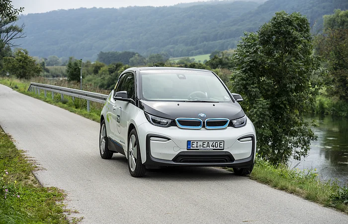Ein weißes Elektroauto fährt auf einer ländlichen Straße entlang eines Flusses, umgeben von Bäumen und Hügeln.