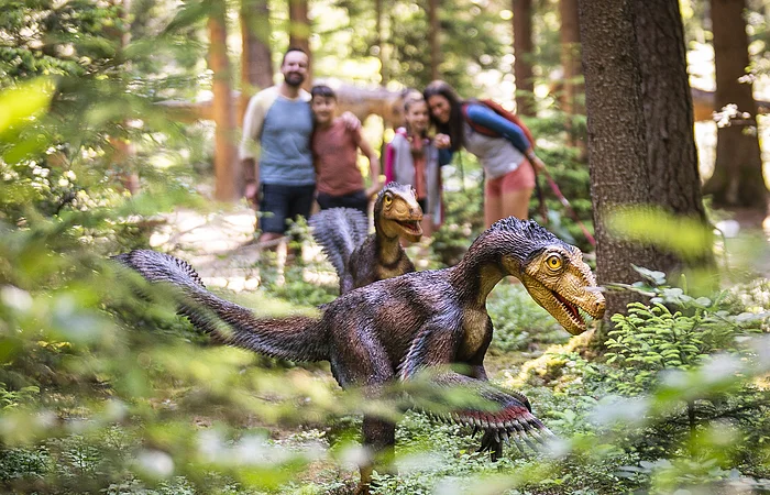 Zwei Dinosauriermodelle im Wald, unscharf im Hintergrund eine Gruppe von Menschen.