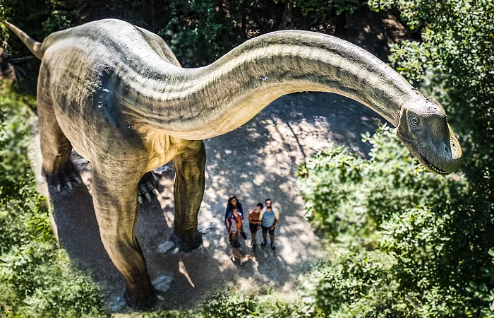 Große Dinosaurierstatue im Wald, Menschen stehen darunter und schauen nach oben.