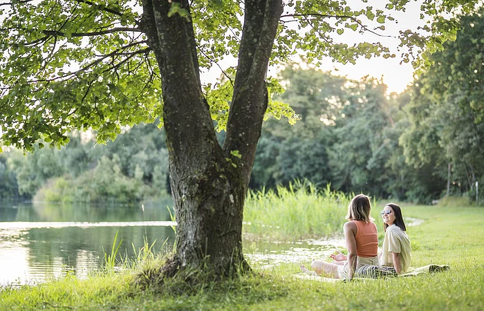 Zwei Frauen liegen auf einer Decke am Leitnerweiher, auch Weicheringer See genannt, und unterhalten sich.
