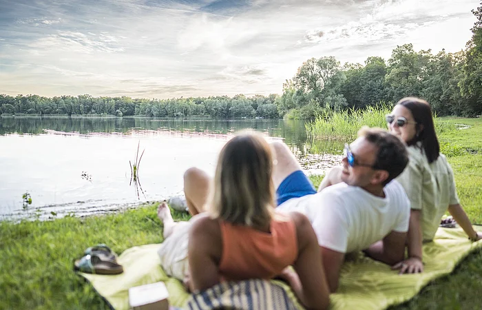 Drei Personen liegen auf einer Decke am Seeufer und schauen auf das Wasser.