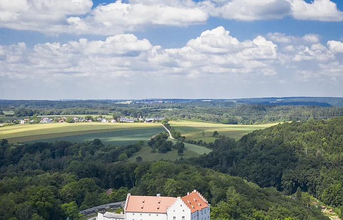 Luftaufnahme eines weißen Schlosses mit rotem Dach, umgeben von Wald und Feldern unter blauem Himmel.