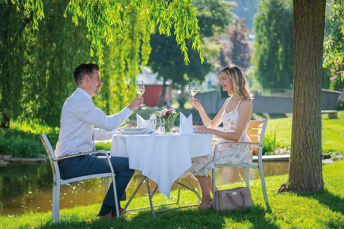 Zwei Personen sitzen an einem Tisch im Freien und stoßen mit Weingläsern an.
