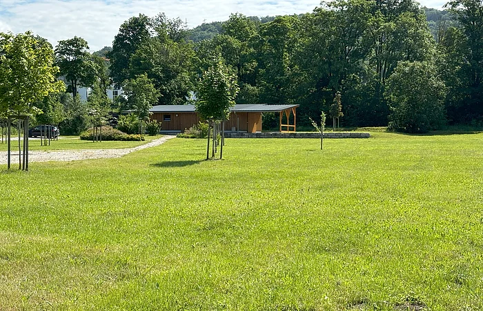 Grüne Wiese mit jungen Bäumen, im Hintergrund ein Holzgebäude und Wald. Ein Kiesweg führt zum Gebäude.