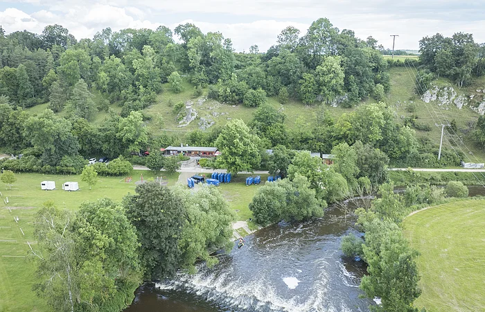 Landschaft mit Fluss, Bäumen, Hügeln und einem Gebäude. Wohnwagen und blaue Boote auf einer Wiese.