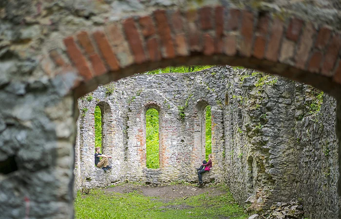Zwei Personen sitzen in den Fensternischen einer alten Steinruine mit grüner Vegetation im Hintergrund.
