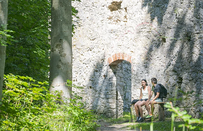 Zwei Personen sitzen auf einer Bank vor einer alten Steinruine im Wald.