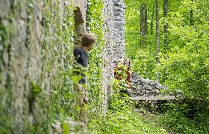 Zwei Personen schauen um eine mit Efeu bewachsene Steinmauer in einem Waldgebiet.