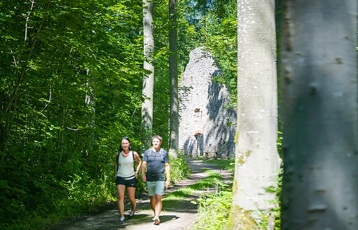 Zwei Personen wandern auf einem Waldweg, im Hintergrund eine alte Steinruine.