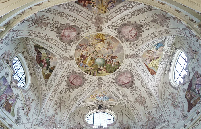 Deckenfresko mit religiösen Szenen und Ornamenten in einer Kirche, umgeben von Fenstern und Stuckverzierungen.