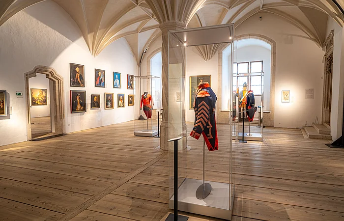 Ein Museumsraum mit Gemälden an den Wänden und historischen Uniformen in Glasvitrinen auf Holzboden.