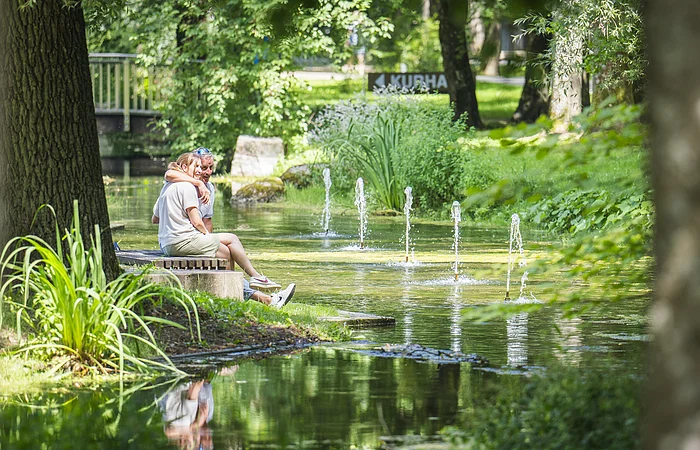 Personen sitzen auf einer Bank am Teich mit Springbrunnen, umgeben von Bäumen und Grünpflanzen.