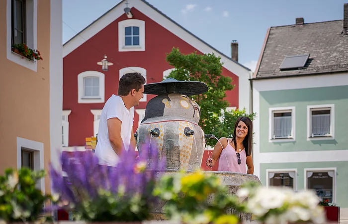 Zwei Personen stehen an einem Brunnen in einer bunten Stadtumgebung.