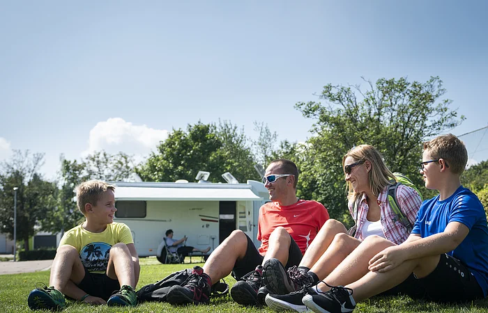 Eine Gruppe von vier Personen sitzt auf einer Wiese vor einem Wohnmobil und lacht.