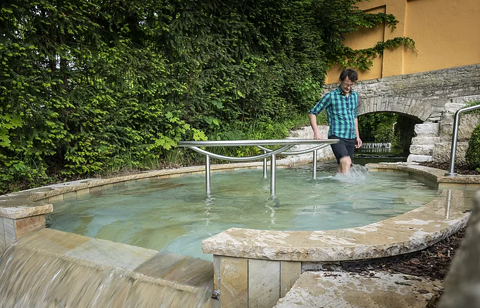 Person im karierten Hemd watet durch ein Kneippbecken mit Handlauf, umgeben von Bäumen und einer Steinmauer.