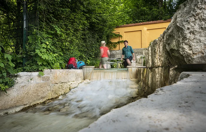 Zwei Personen gehen an einem kleinen Wasserlauf vorbei, umgeben von grüner Vegetation und Steinmauern.