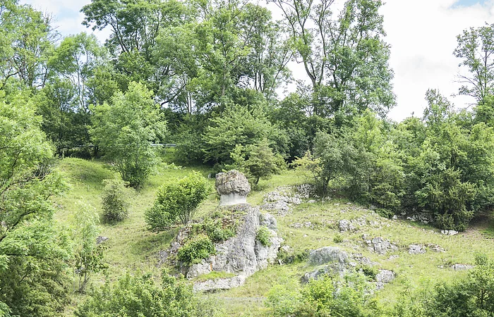 Grüne Hügellandschaft mit Felsen, Bäumen und einem Gebäude im Vordergrund. Spielplatz mit Klettergerüst sichtbar.