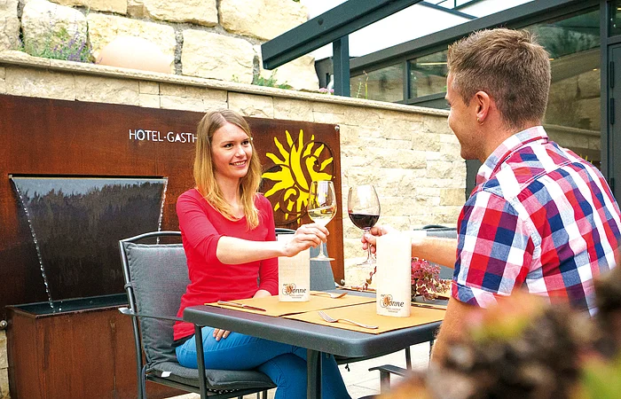 Zwei Personen sitzen an einem Tisch im Freien, stoßen mit Weingläsern an, im Hintergrund eine Steinmauer.