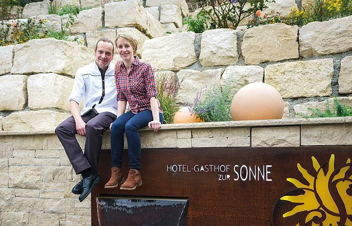 Zwei Personen sitzen auf einer Steinmauer neben einem Schild mit der Aufschrift "Hotel-Gasthof zur Sonne".