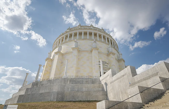 Rundes, klassizistisches Gebäude mit Säulen und Statuen, umgeben von Treppen, unter blauem Himmel mit Wolken.