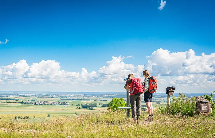 Zwei Personen mit Rucksäcken stehen auf einem Hügel und blicken durch ein Fernglas auf eine weite Landschaft.