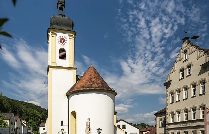 Kirche mit Turm und Uhr, umgeben von Gebäuden und Bäumen, unter blauem Himmel mit Wolken.