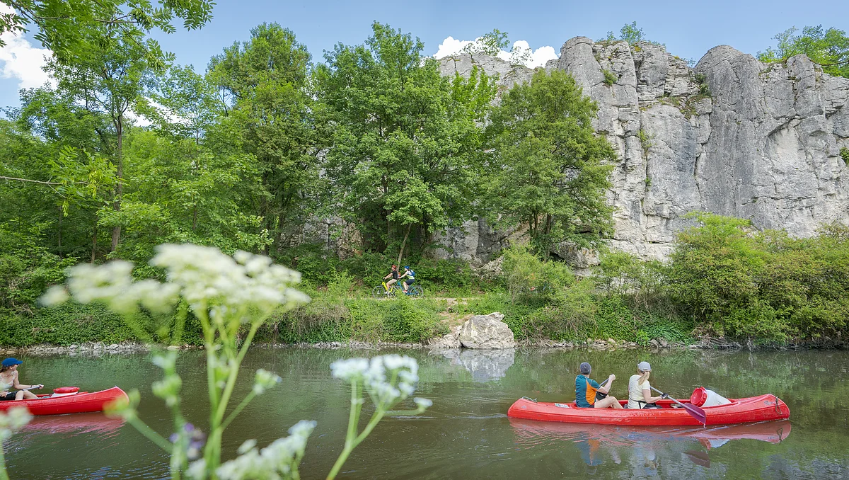 Zwei rote Kanus auf einem Fluss, im Hintergrund Felsen und Bäume, im Vordergrund unscharfe Blumen.