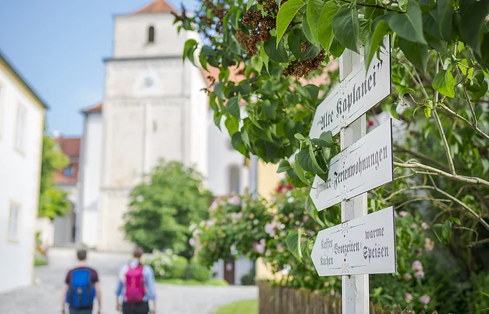 Zwei Personen gehen auf einem gepflasterten Weg, im Hintergrund eine Kirche. Ein Wegweiser steht rechts.