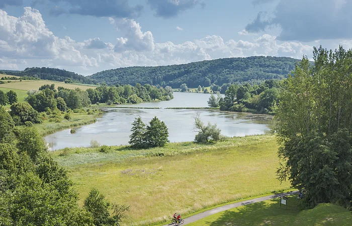 Landschaft mit Fluss, Wiesen und bewaldeten Hügeln im Hintergrund, zwei Radfahrer auf einem Weg im Vordergrund.