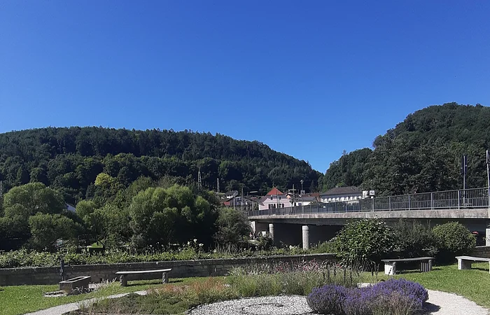 Garten mit Kieswegen und Pflanzen, im Hintergrund eine Brücke und bewaldete Hügel unter blauem Himmel.