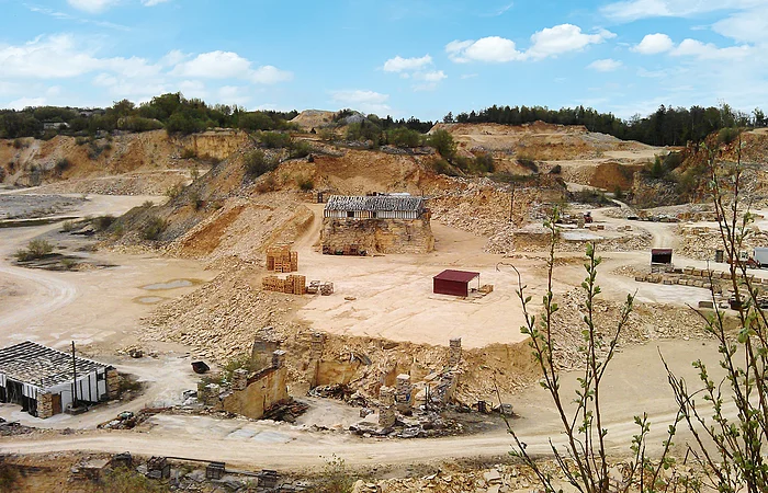 Steinbruch mit Sand- und Felsformationen, Gebäuderuinen und Paletten, umgeben von Vegetation.