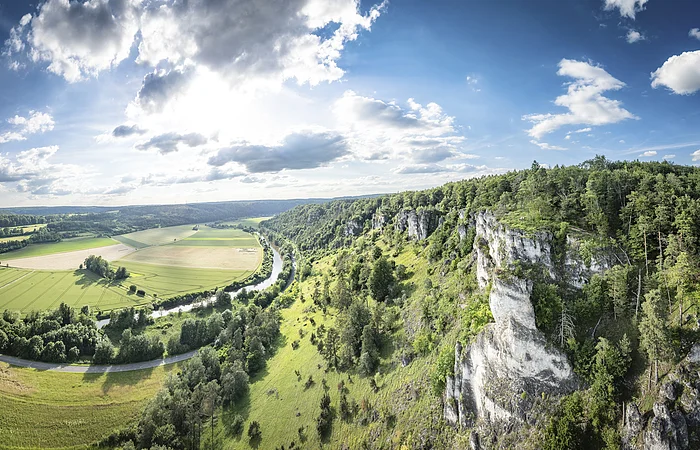 Landschaft mit grünen Feldern, einem Fluss und bewaldeten Hügeln unter einem bewölkten Himmel.