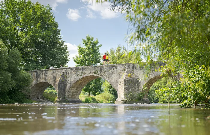 Steinbrücke mit drei Bögen über einen Fluss, umgeben von Bäumen und Vegetation.
