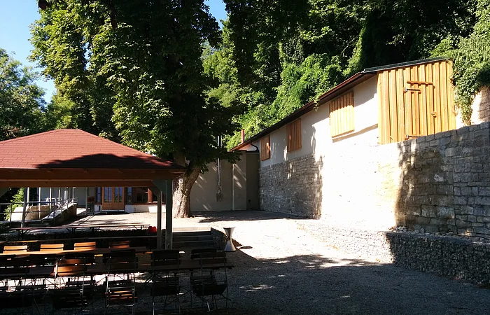 Außenbereich mit Holztischen und -stühlen, überdachtem Pavillon, Steinmauer und Bäumen im Hintergrund.