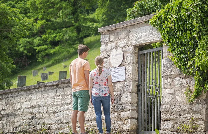 Ein Mann und eine Frau stehen vor einem Eingangstor mit einem Davidstern, umgeben von einer Steinmauer und Bäumen.