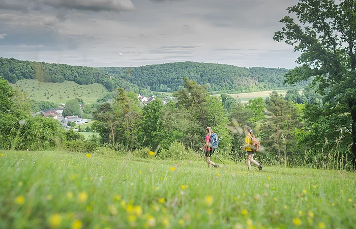 Zwei Personen wandern über eine Wiese mit Blick auf ein Dorf und bewaldete Hügel im Hintergrund.
