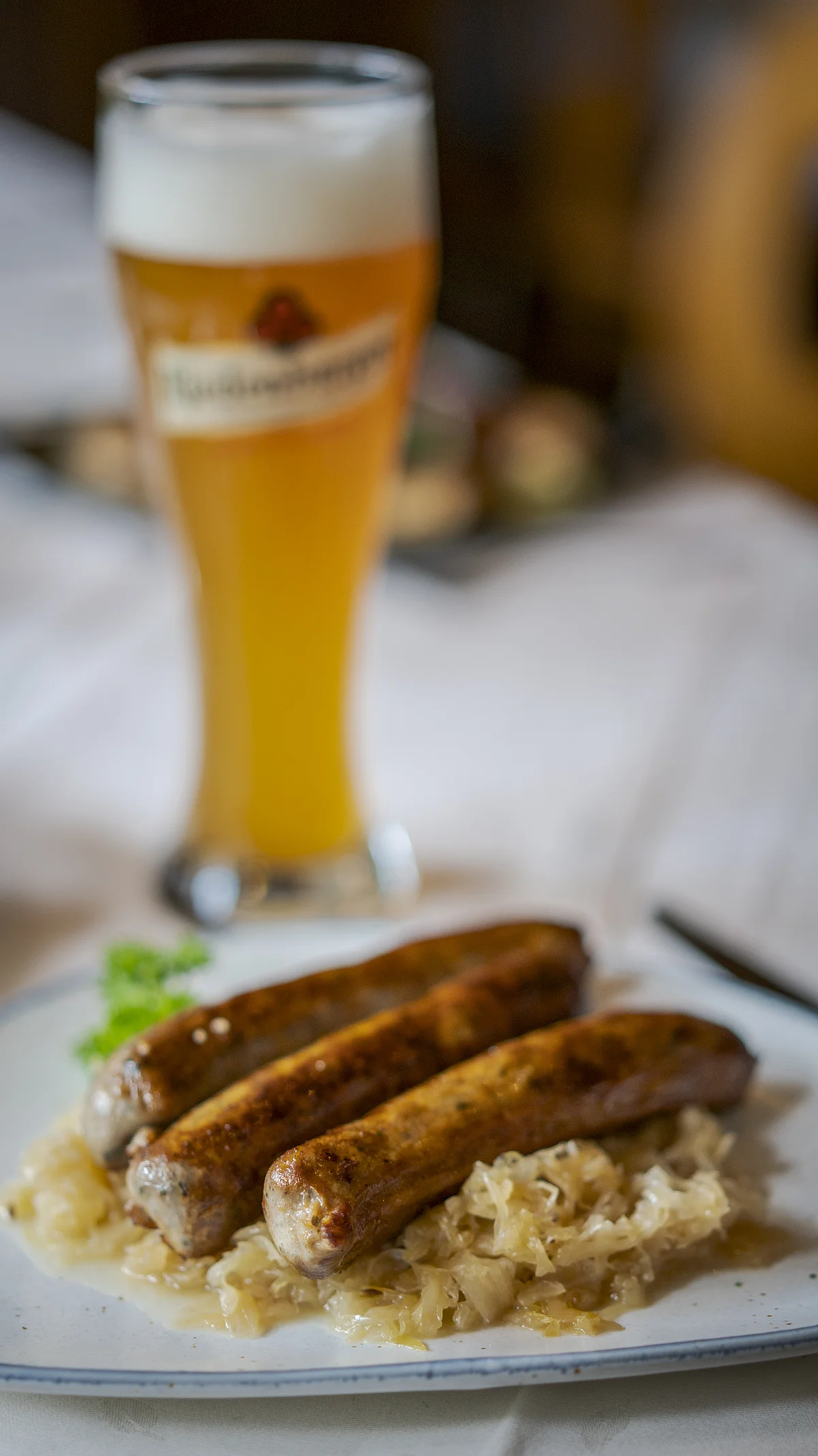 Drei Bratwürste auf Sauerkraut auf einem Teller, im Hintergrund ein Weizenglas mit Bier gefüllt.