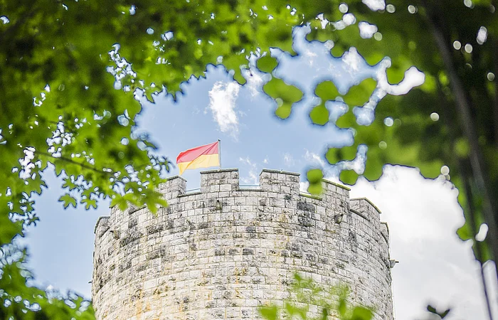 Ein runder Steinturm mit Zinnen und einer rot-gelben Flagge, umrahmt von grünen Blättern.