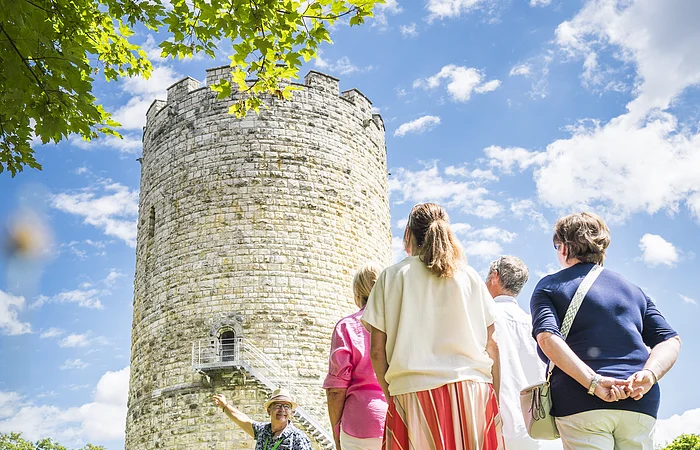 Gruppe von Menschen steht vor einem alten Steinturm, ein Mann zeigt auf den Turm.