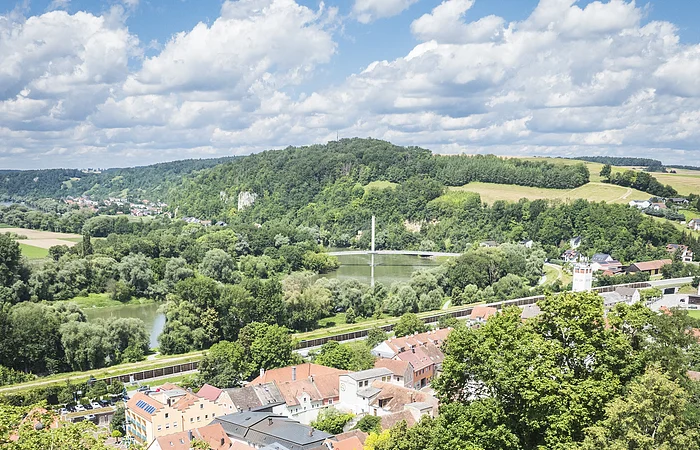 Landschaft mit Fluss, Brücke, Hügeln und einer Stadt mit roten Dächern, umgeben von Bäumen und Wiesen.